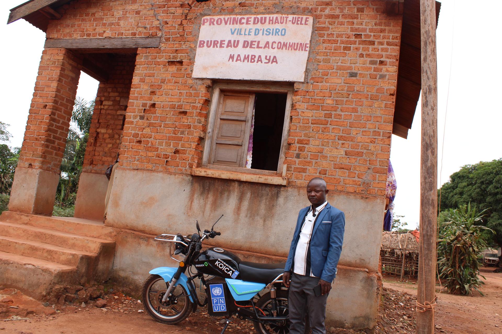 Photograph: man in a denim jacket beside a blue motorcycle in front of a small brick building.