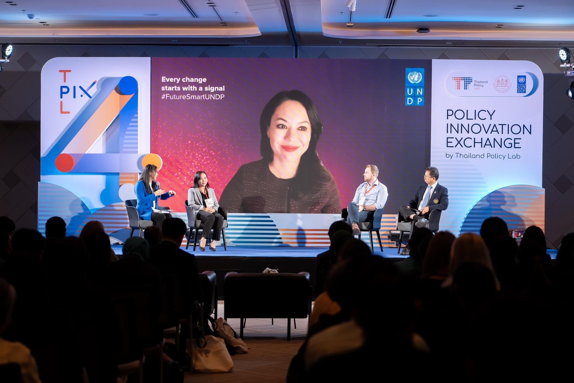 Panelists on stage at a conference; large screen displays a blurred speaker, audience silhouettes in foreground.