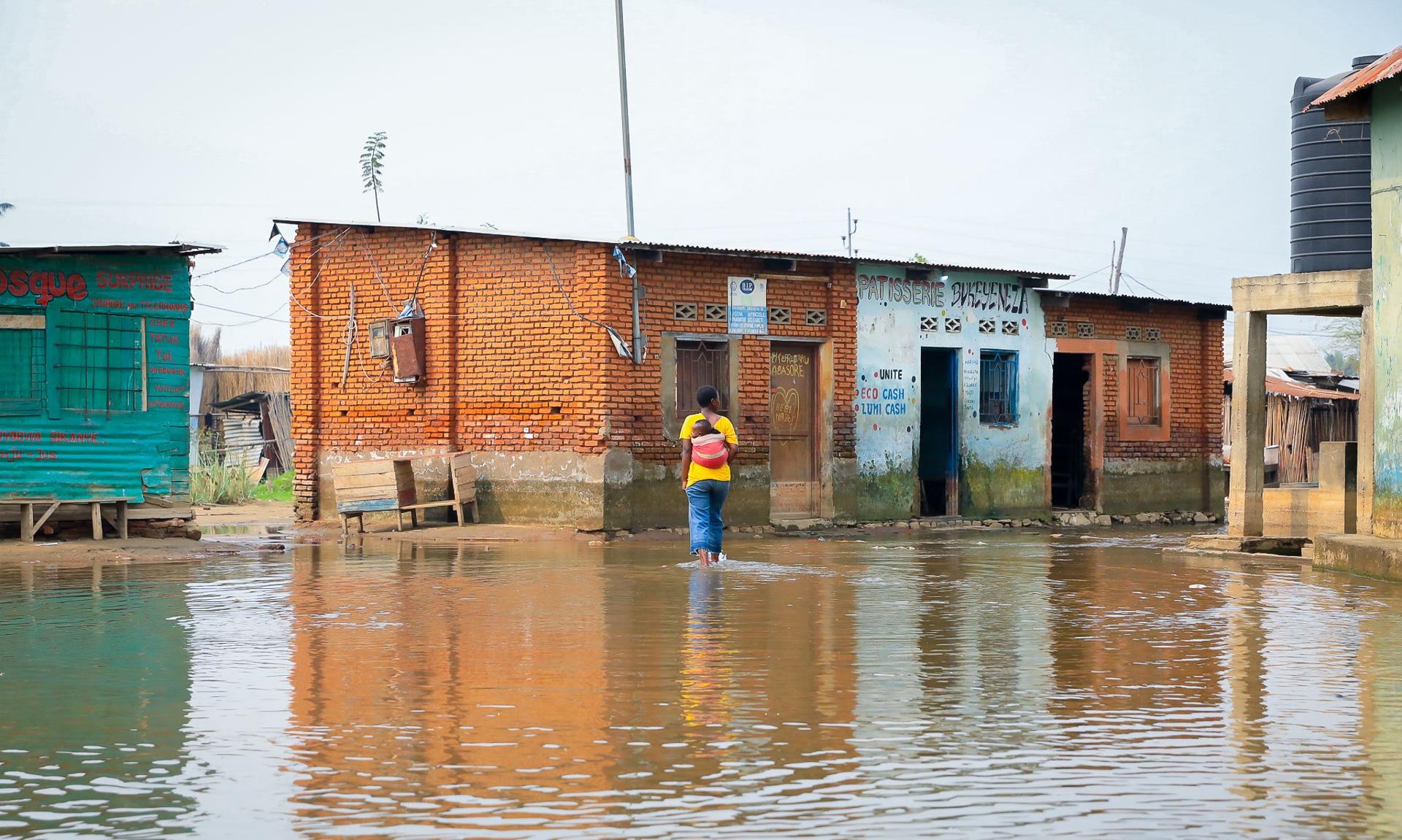 Person in bright clothing wades through floodwaters on a colorful street with submerged houses.