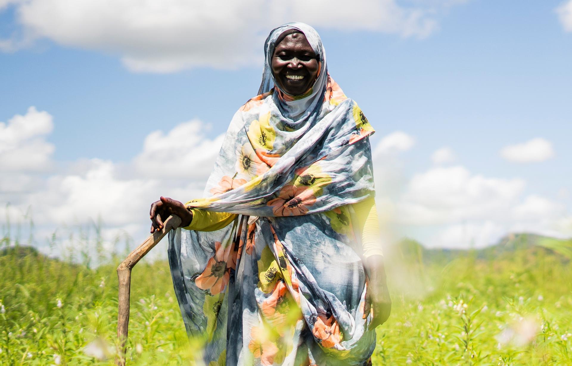 Smiling person in a colorful shawl and sunglasses, holding a wooden staff in a sunny grassy field.