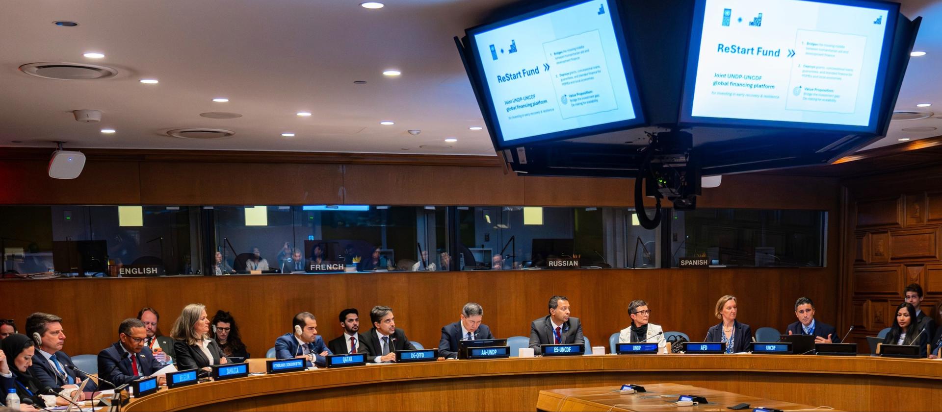 Photograph of attendees in a boardroom around a long conference table with overhead monitors.