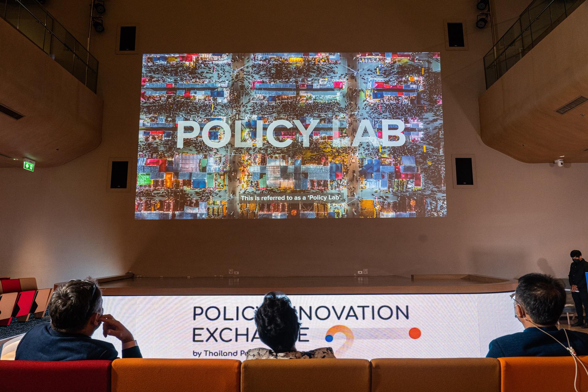 Panelists on stage in a conference hall; mosaic collage screen behind displaying POLICY-LAB.
