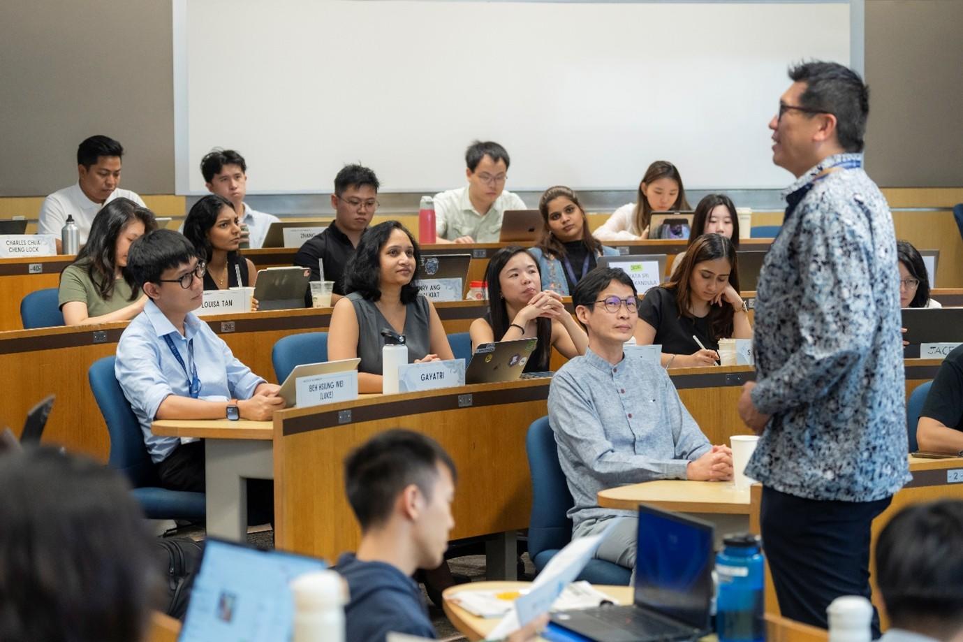 Photograph: lecturer in a patterned shirt standing at the front of a classroom full of students.