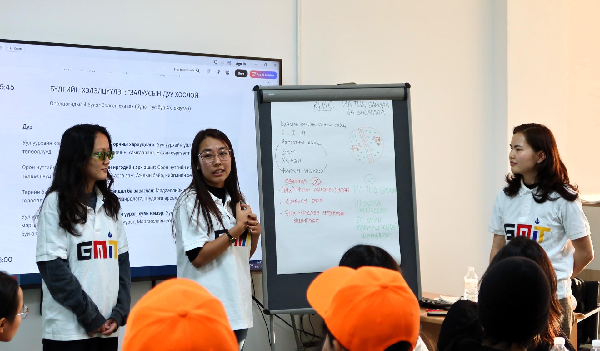 Photo: three women presenters in a classroom with screen and flip chart; audience in orange hats.