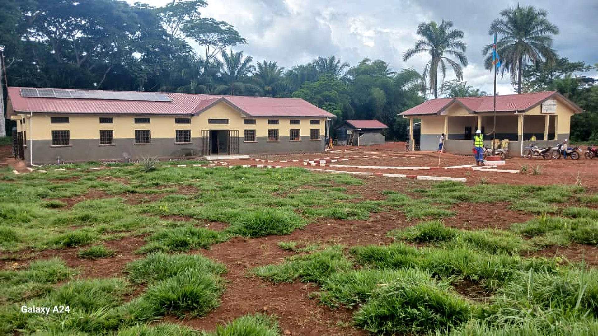 Two single-story beige buildings with red roofs; bare brown ground and green shrubs and palm trees.