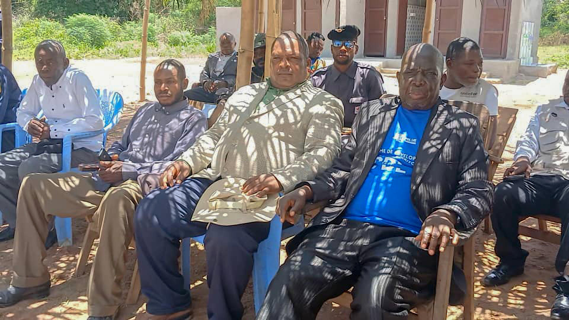 Group of adults seated on plastic chairs outdoors on a sunny day.