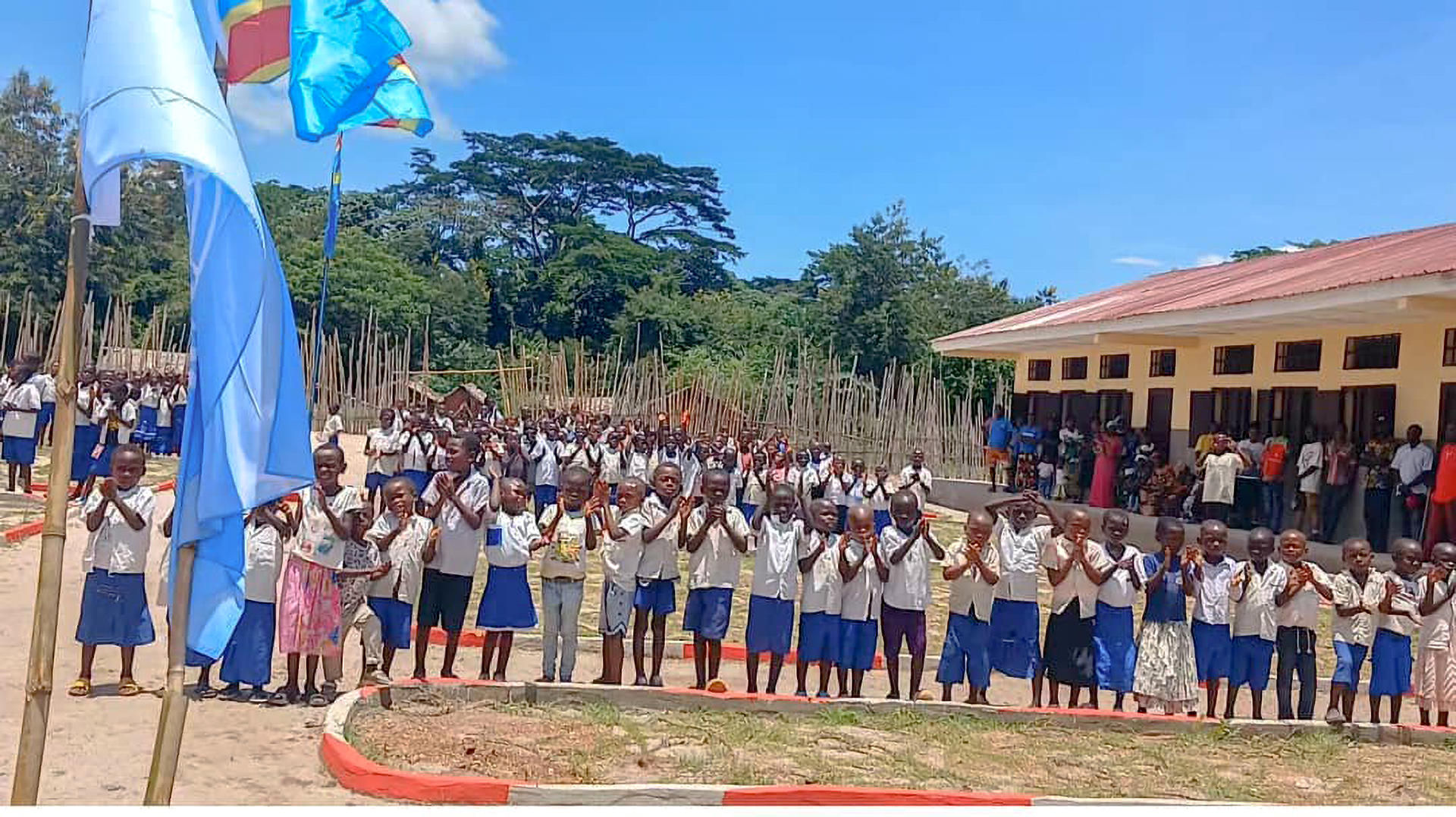 Group of students lined up outdoors on a sandy area, yellow building to the right, blue flags left.