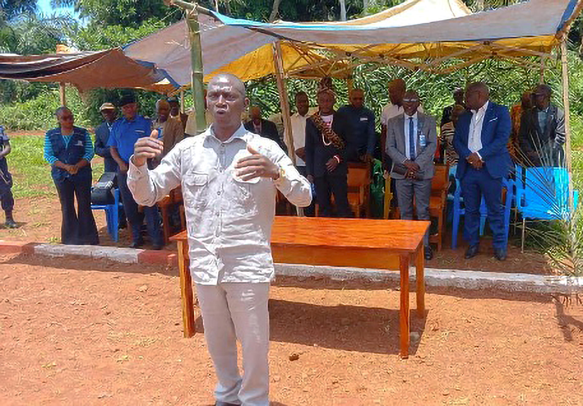 Man in light clothing addresses a gathered crowd under yellow canopies outdoors.
