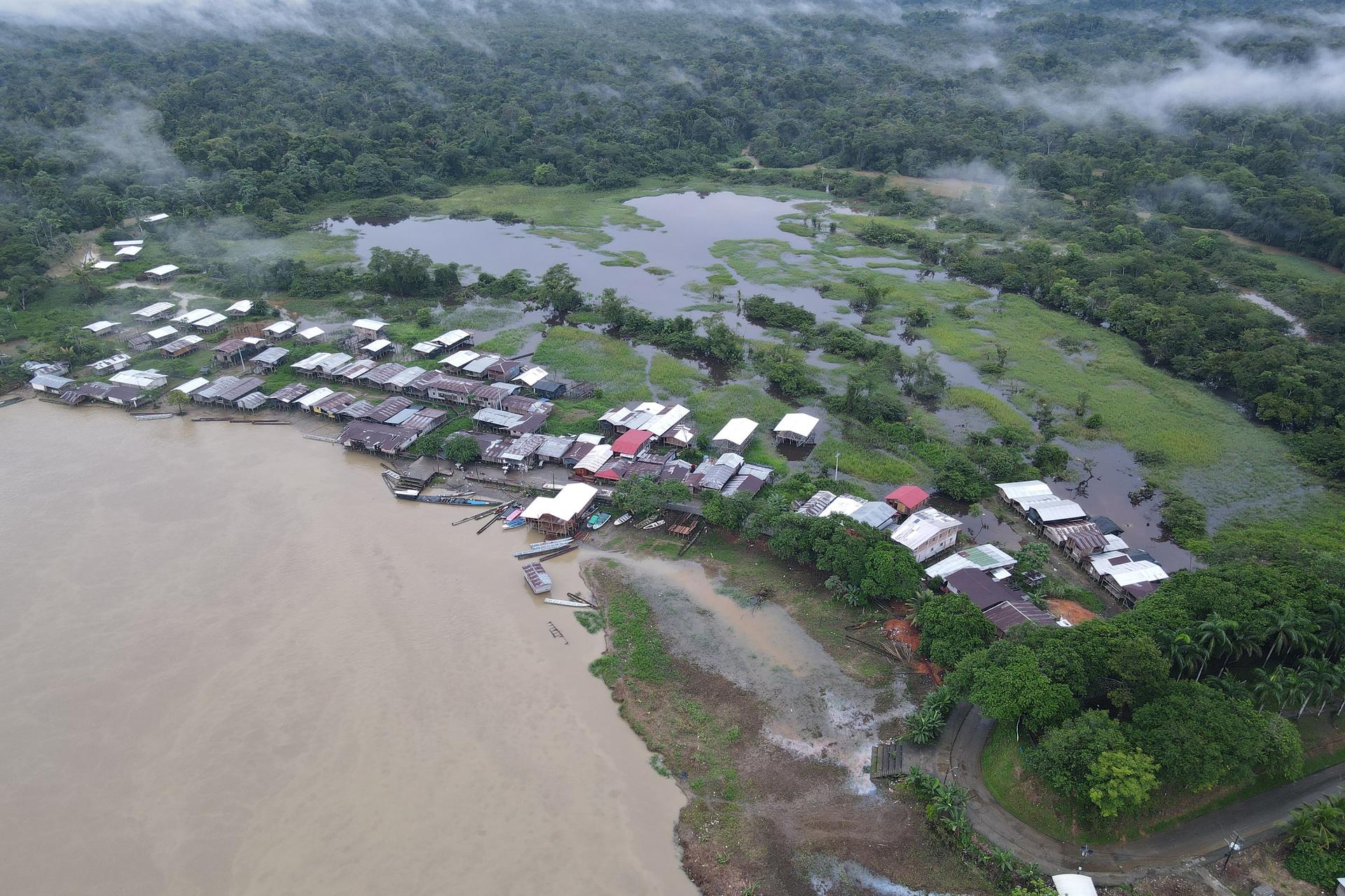 Aerial view of a riverside village with stilt houses along a muddy shoreline amid green fields.
