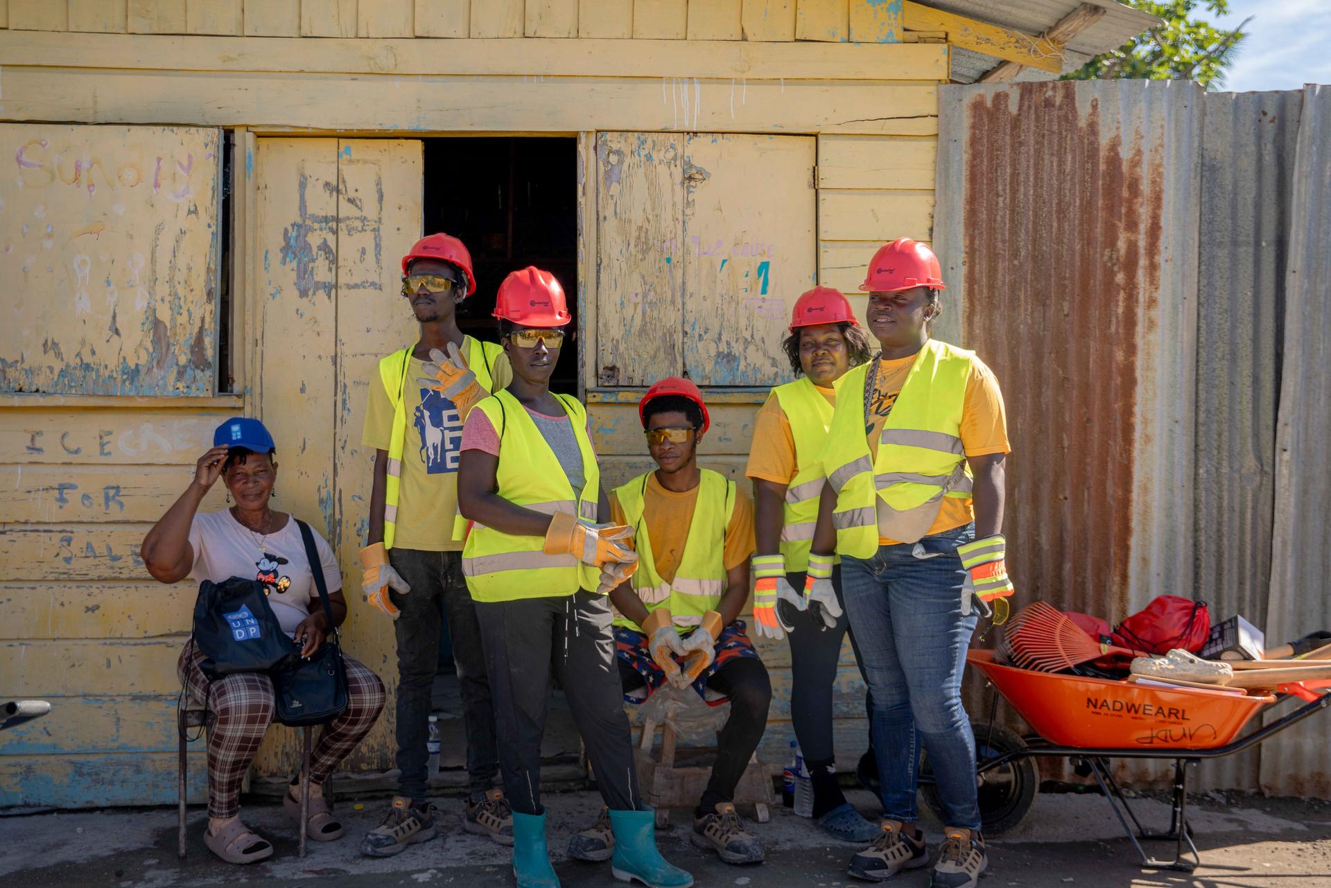 Group of construction workers in hard hats and high-visibility vests pose outside a wooden construction site.
