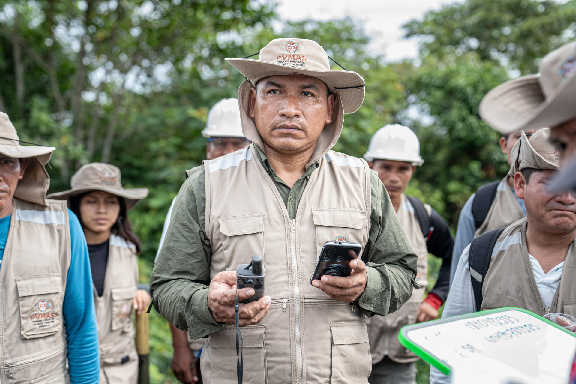 Outdoor tour group wearing wide-brim hats; central man in beige shirt holds two smartphones.