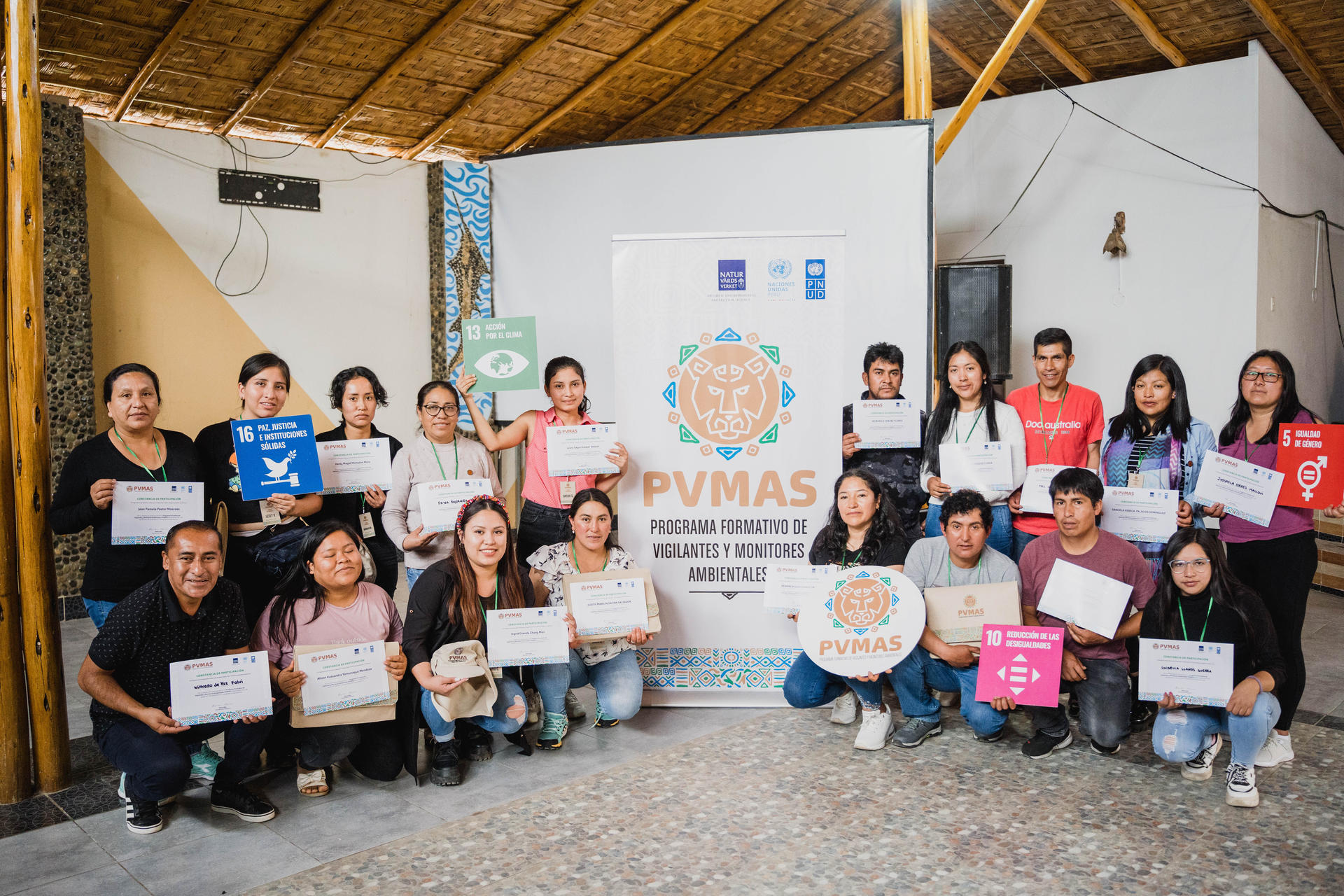 Group photo of workshop participants posing with signs under a banner in a thatched shelter.