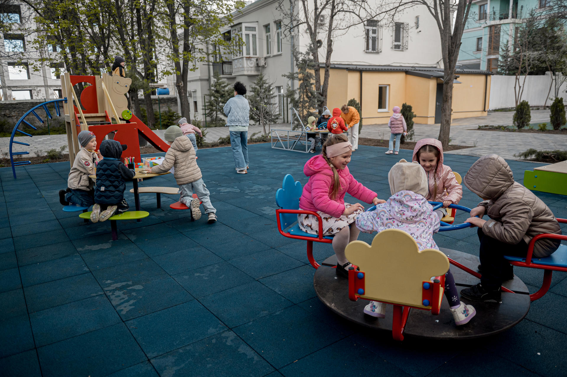 Kids sit at round car-shaped tables in a blue playground while adults watch.