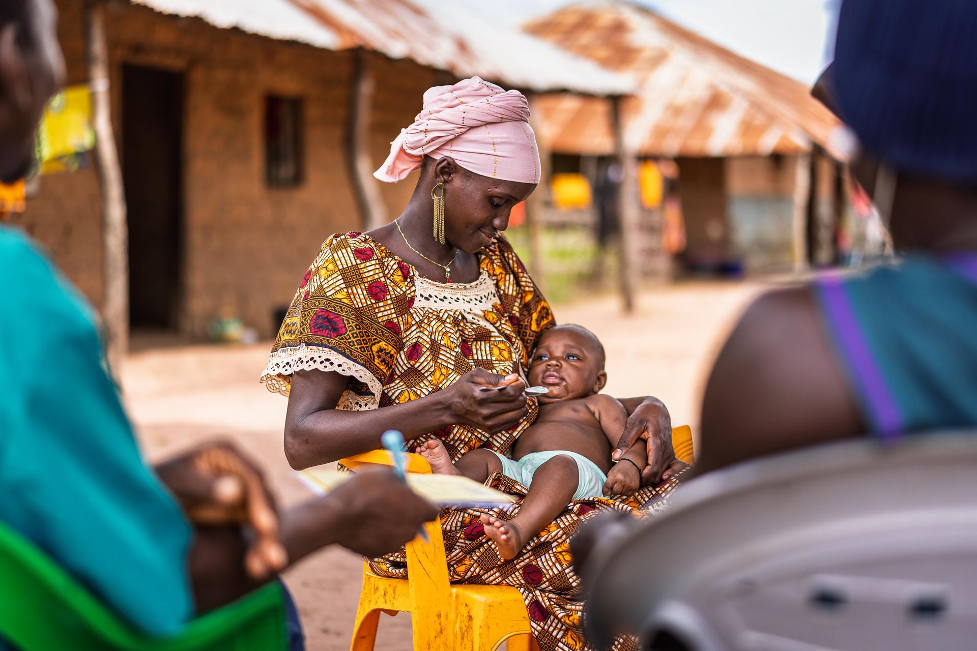Photograph of a woman in a patterned dress holding a baby, in a sunny village with thatched huts.