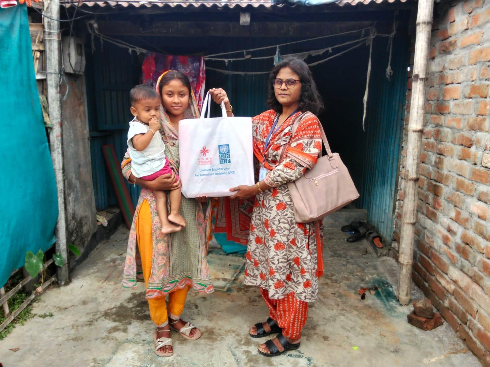 Two women with a baby stand outside a brick doorway; one holds a framed certificate.
