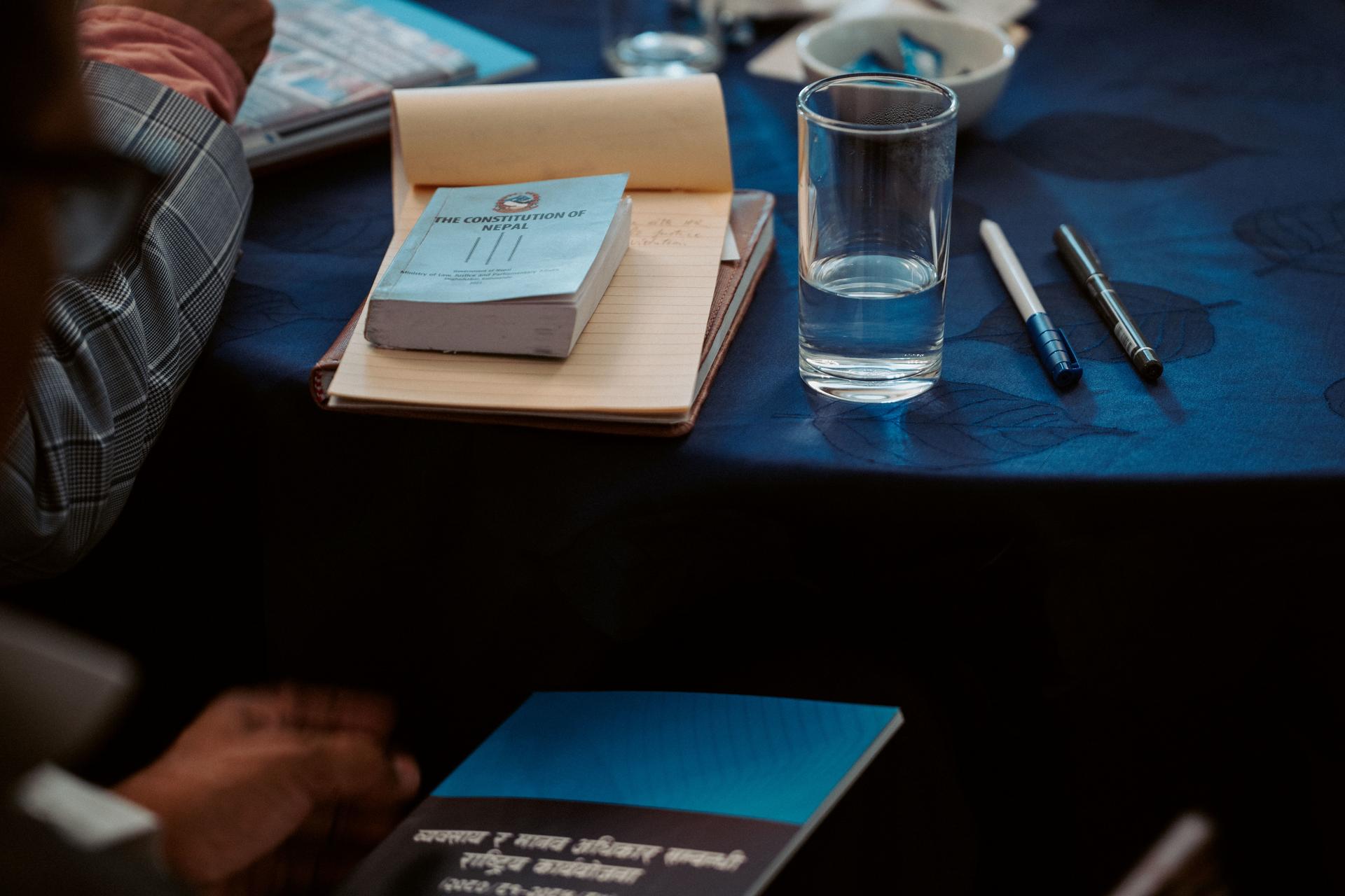 Close-up of a blue tablecloth with a glass of water, pens, and notebooks.
