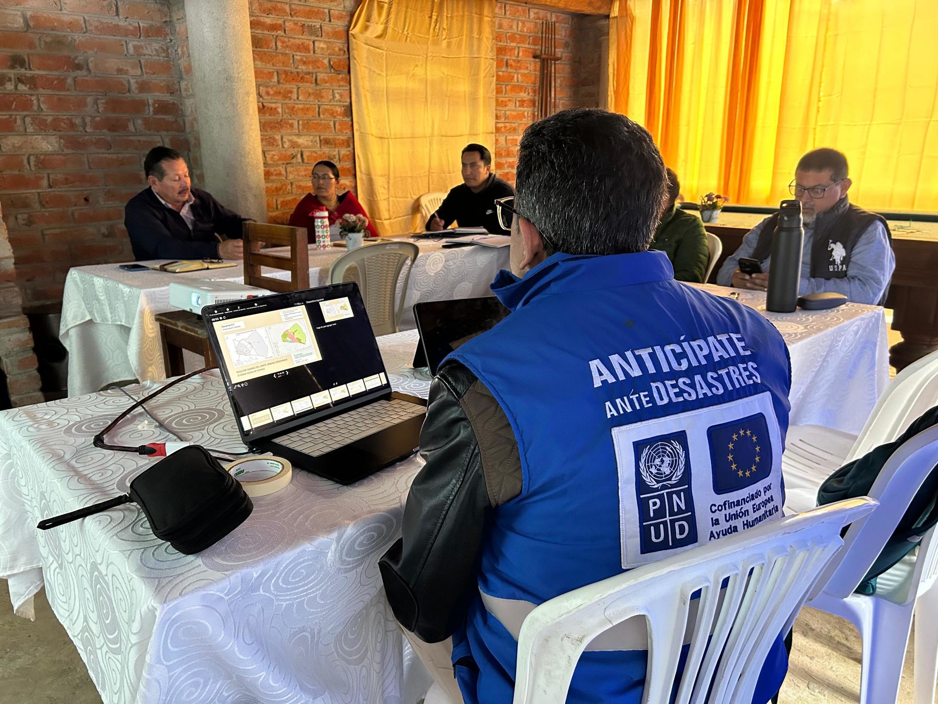 Group of people in a training room; man in a blue vest with white text on back sits at a laptop.