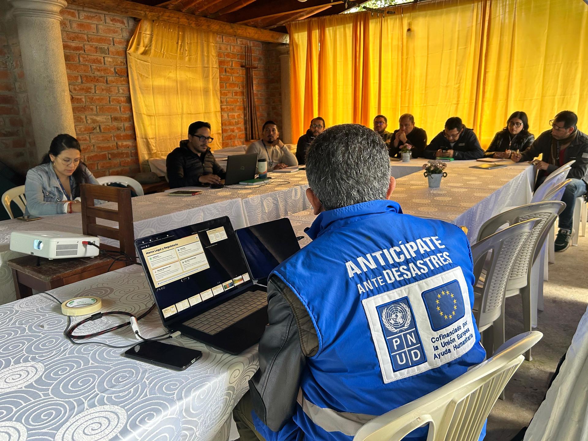 Group of attendees around tables; foreground person in blue vest faces away at a laptop.