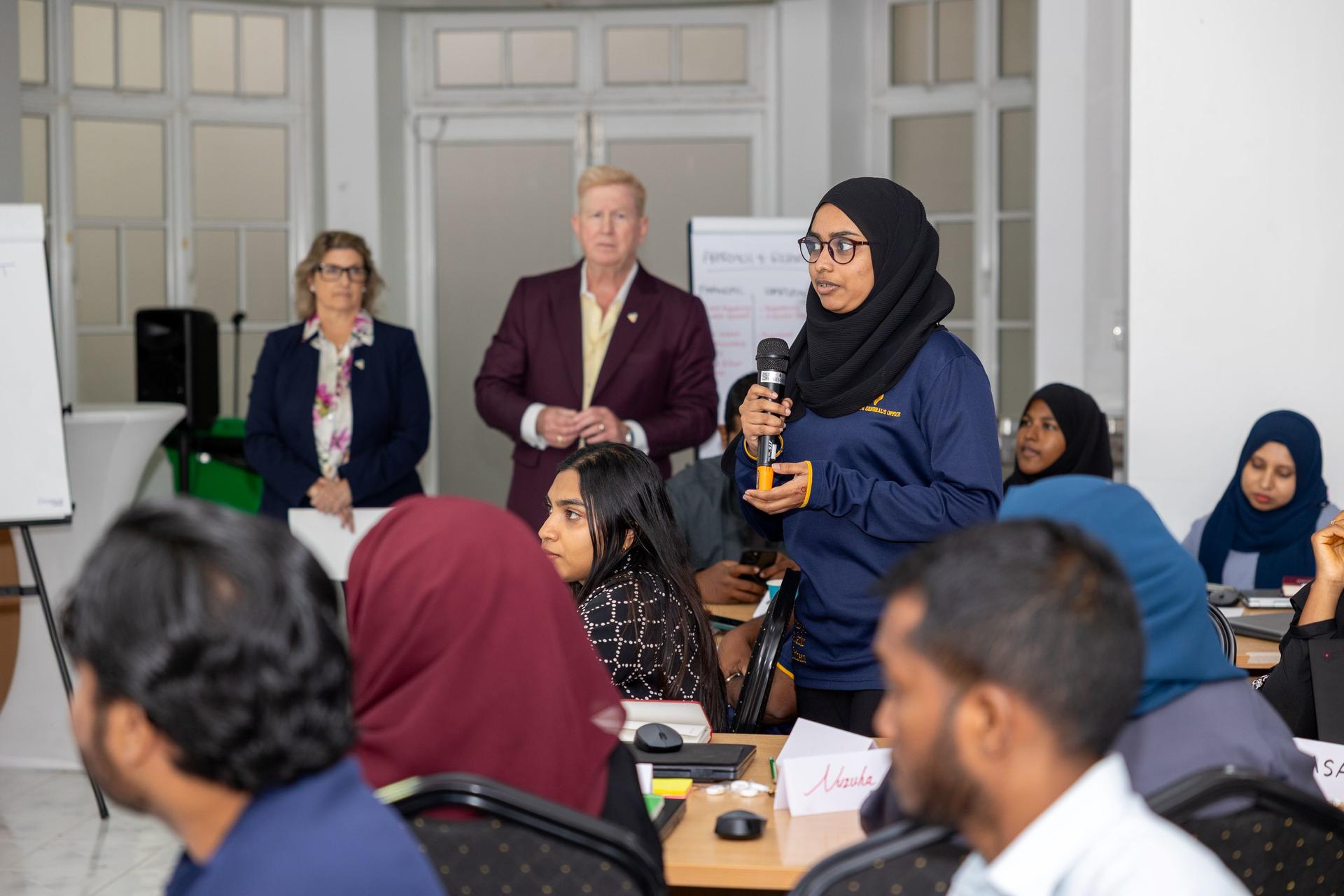 In a conference room, a woman wearing a hijab addresses a seated, diverse audience.