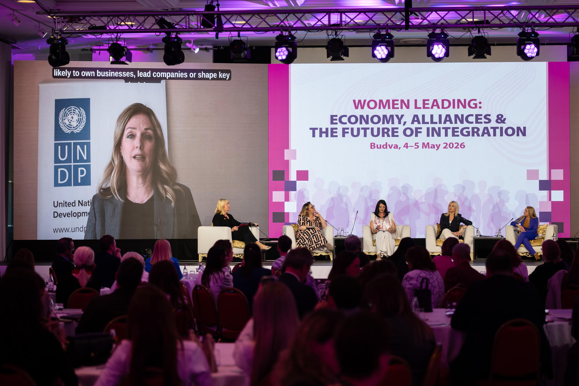Conference stage with female panelists, a large video screen, and a purple banner backdrop.