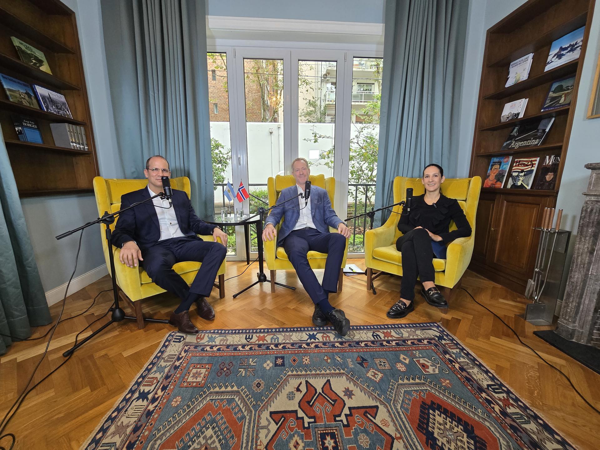 Photograph of three smiling men in suits sitting on colorful chairs in a bright living room.