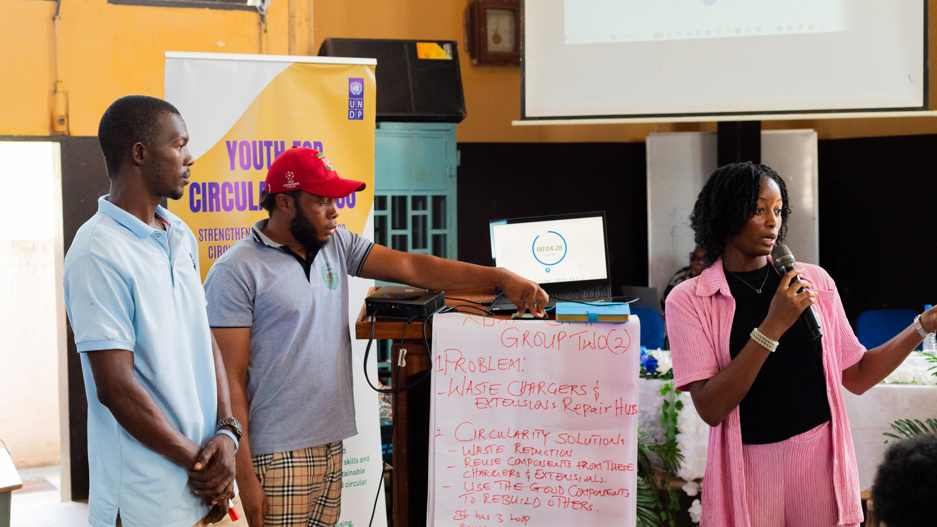 Group of people presenting on a stage with a pink signboard and projector screen in the background.