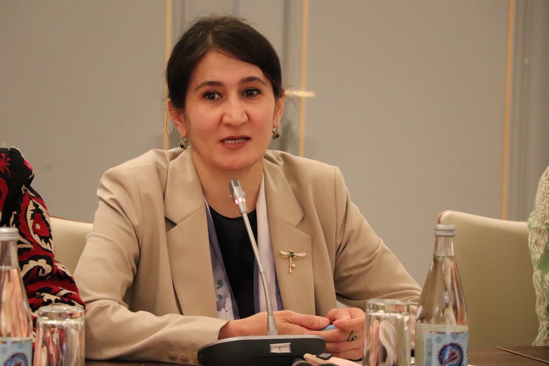 Blurry-faced person in beige blazer seated at conference table with water bottles, a microphone, and a phone.
