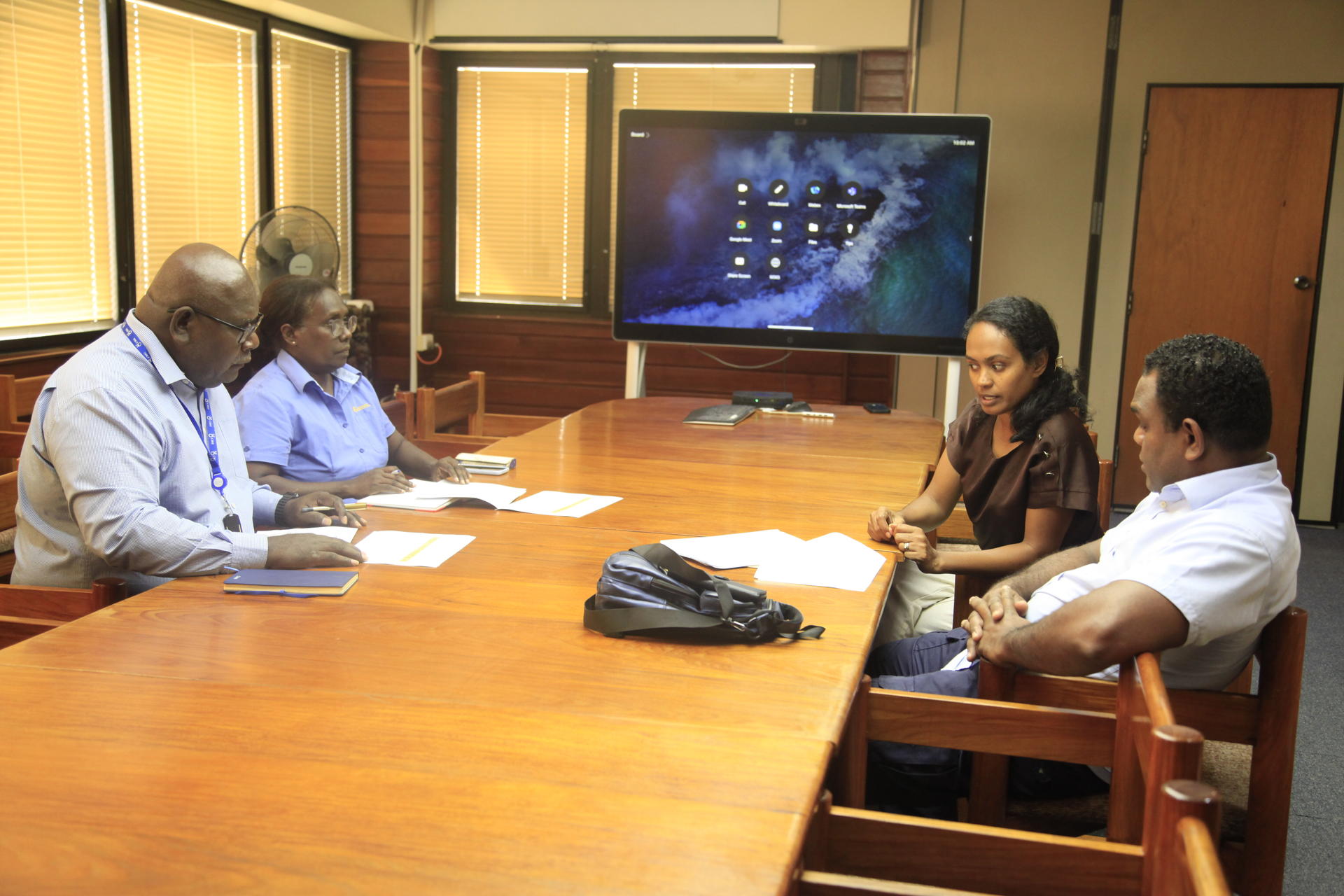Photo: group of people seated around a long wooden conference table in a meeting room.