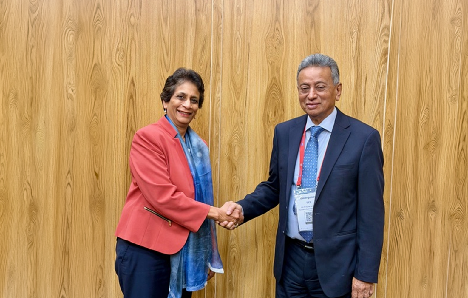 Two professionals shake hands in front of a wood wall; woman in pink blazer, blue top, man in suit.