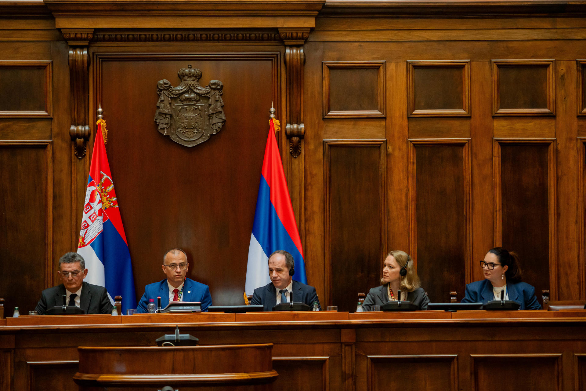 Five officials seated at a wood-paneled dais with microphones, flags displayed behind.