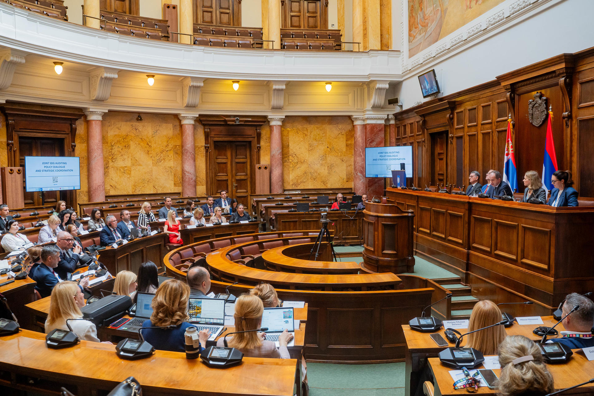 Photo of a large wood-paneled parliamentary chamber with a speaker at a podium and an audience.