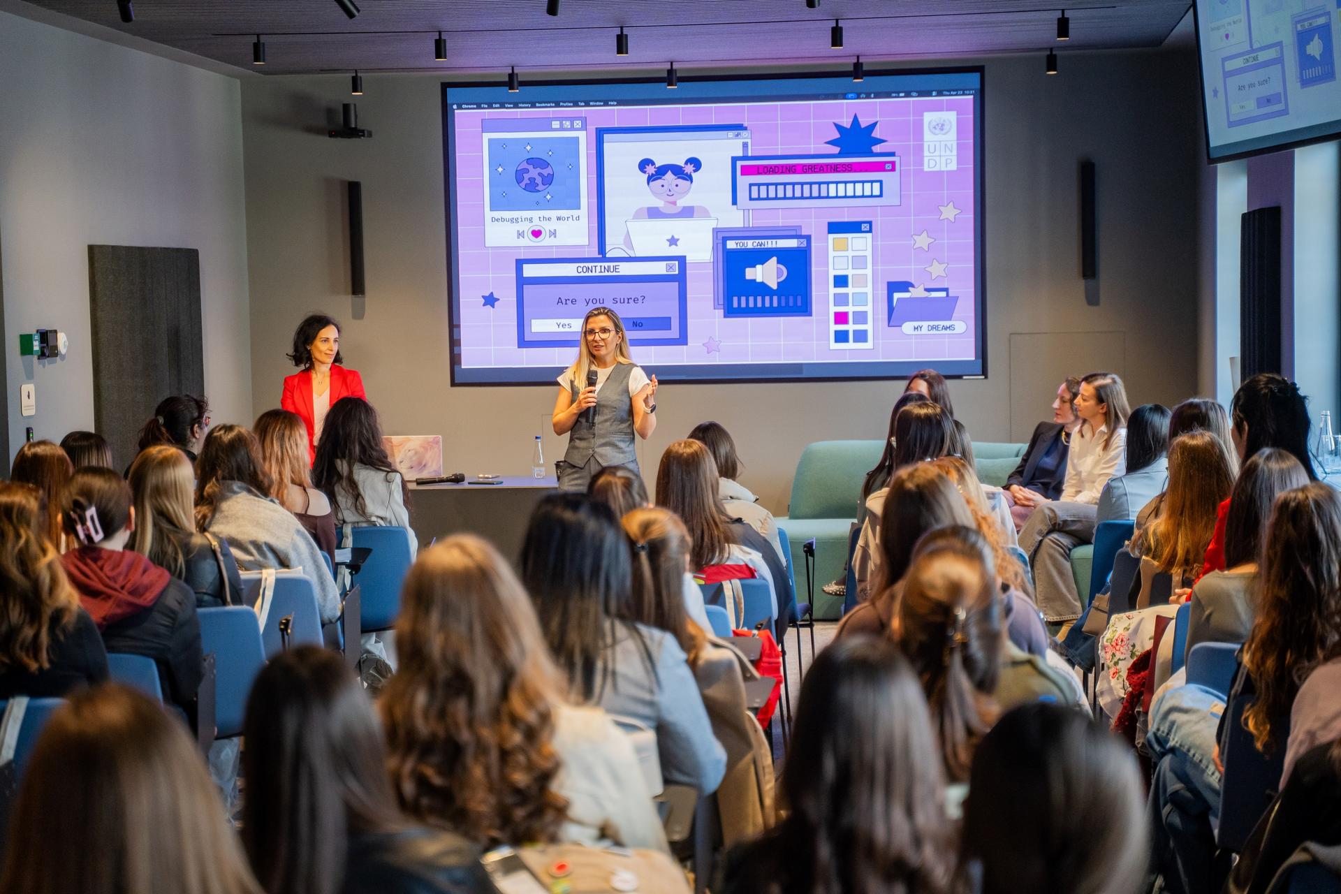 Photograph of a conference room with a presenter onstage, seated audience, and slides projected.