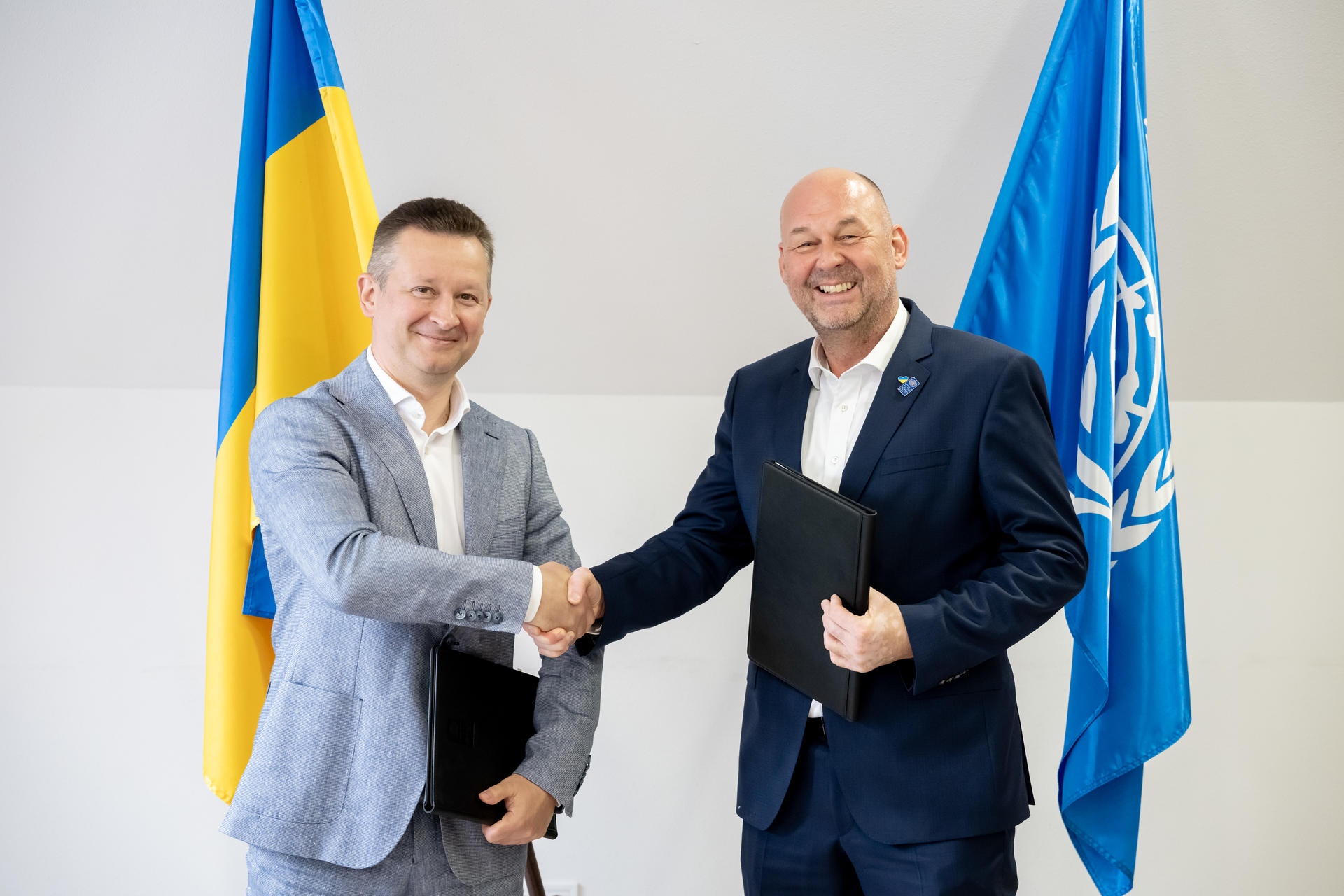 Two men in suits shake hands in front of Ukrainian and UN flags; one holds a document.