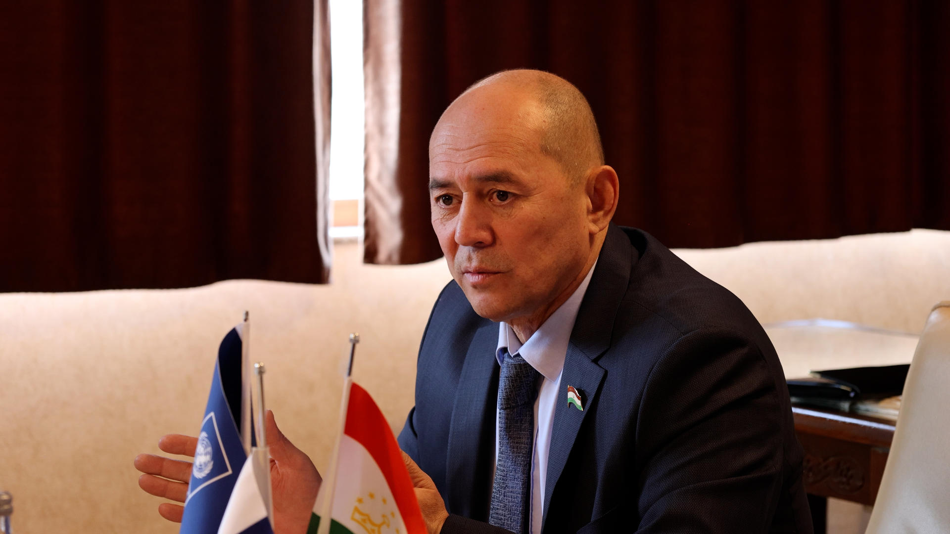 Photograph of a suited man seated at a table with flags in the background; face blurred.