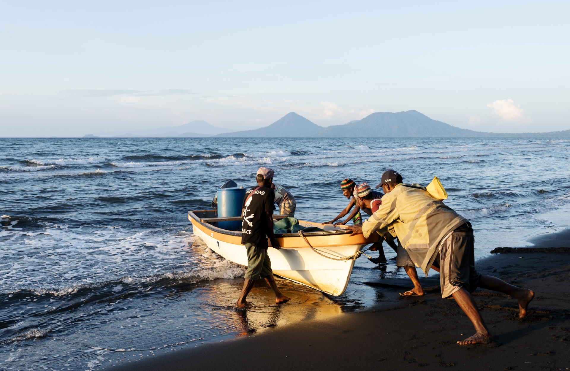 Photograph of three people launching a small boat from a sandy shore into the blue sea.