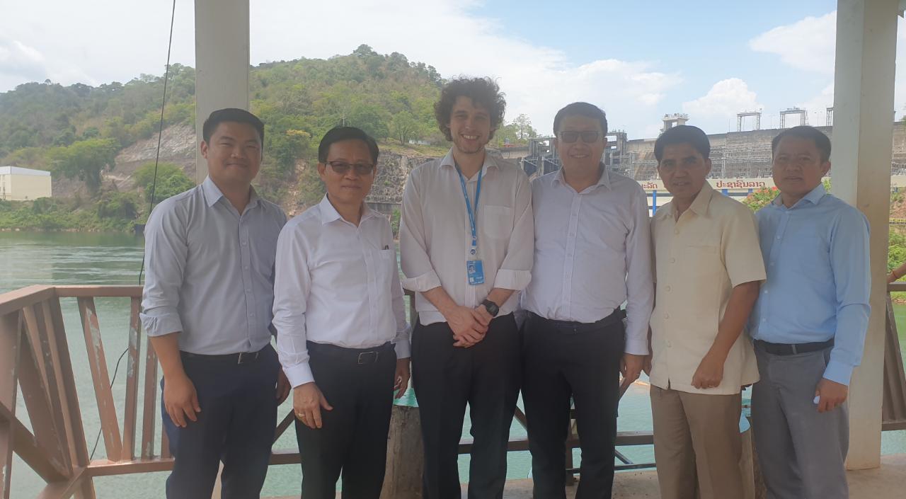 Group of men in business attire posing on an outdoor waterfront terrace with hills in background.