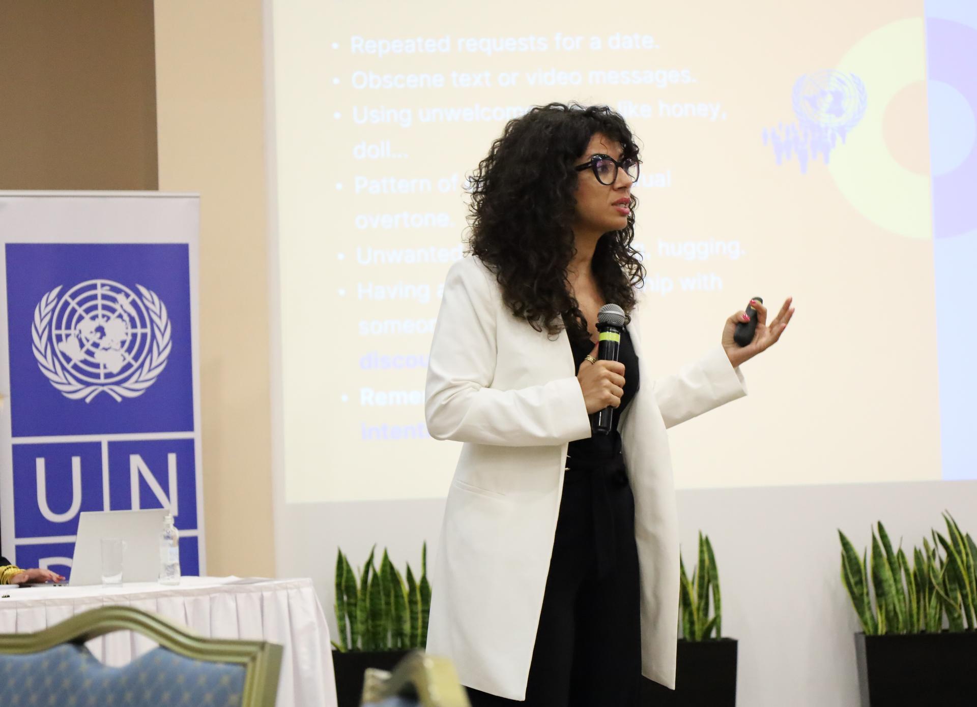 Female presenter in a white blazer speaks into a microphone on stage beside a UN banner.