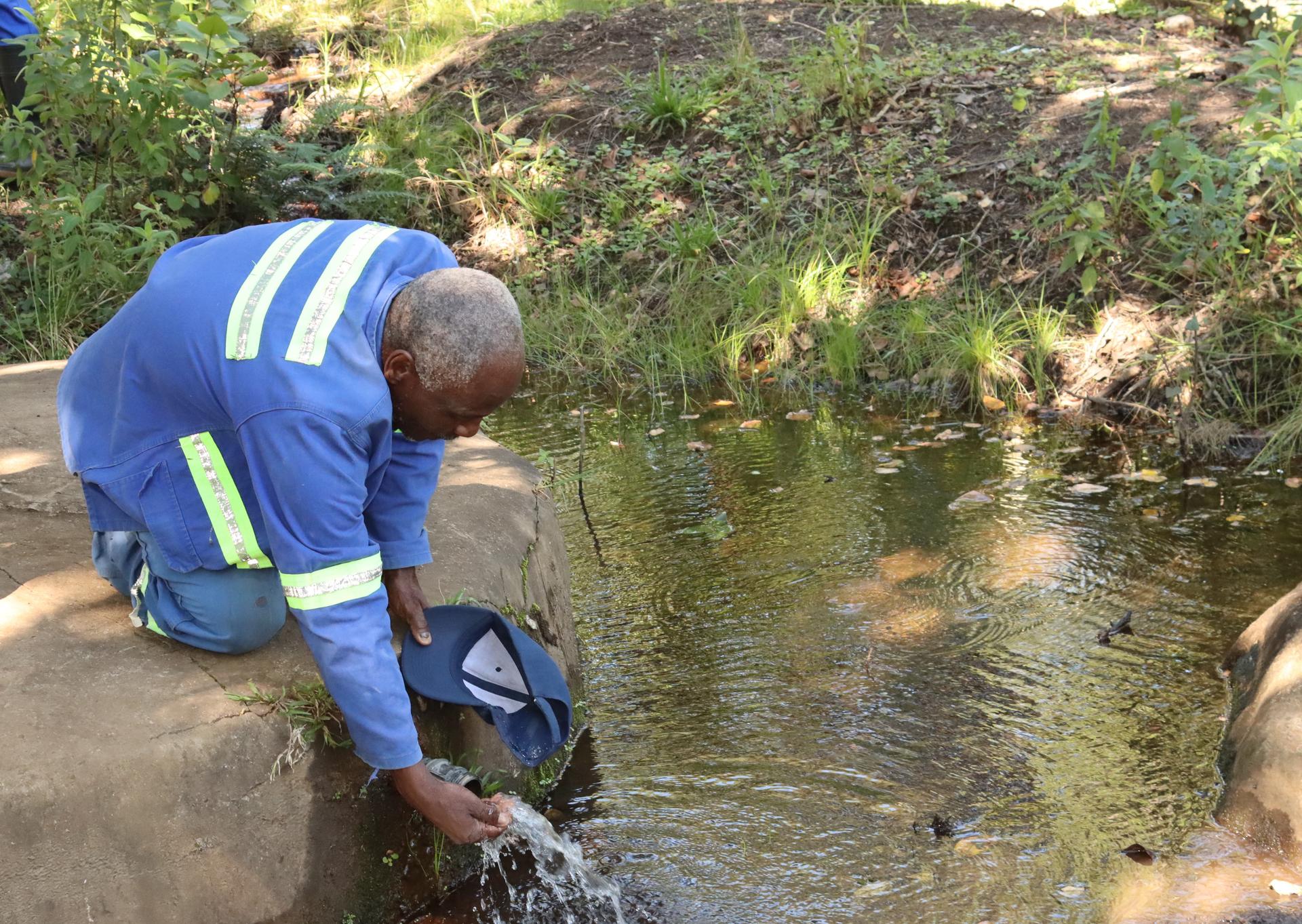 A man in blue with reflective stripes crouches by a creek, dipping a container into water.