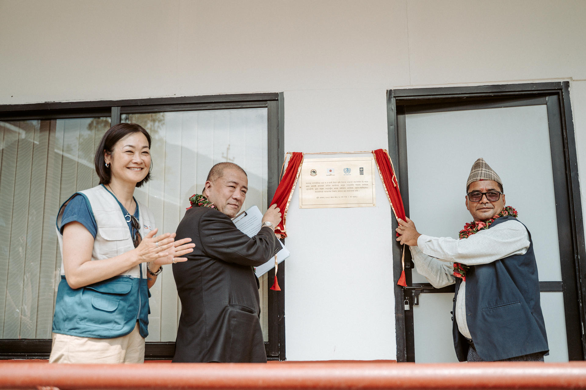 Three people on a balcony at a ribbon-cutting ceremony; center man cuts a red ribbon.