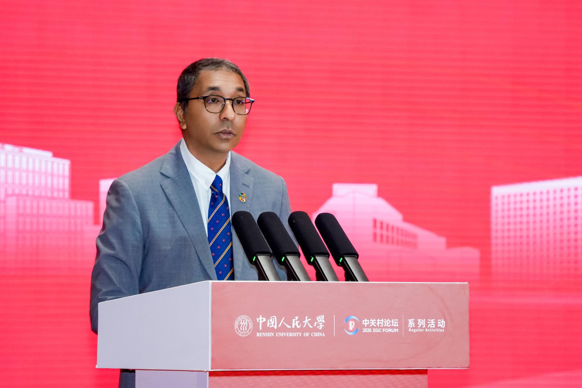 Man in gray suit and blue tie speaks at a podium with microphones on a red backdrop.