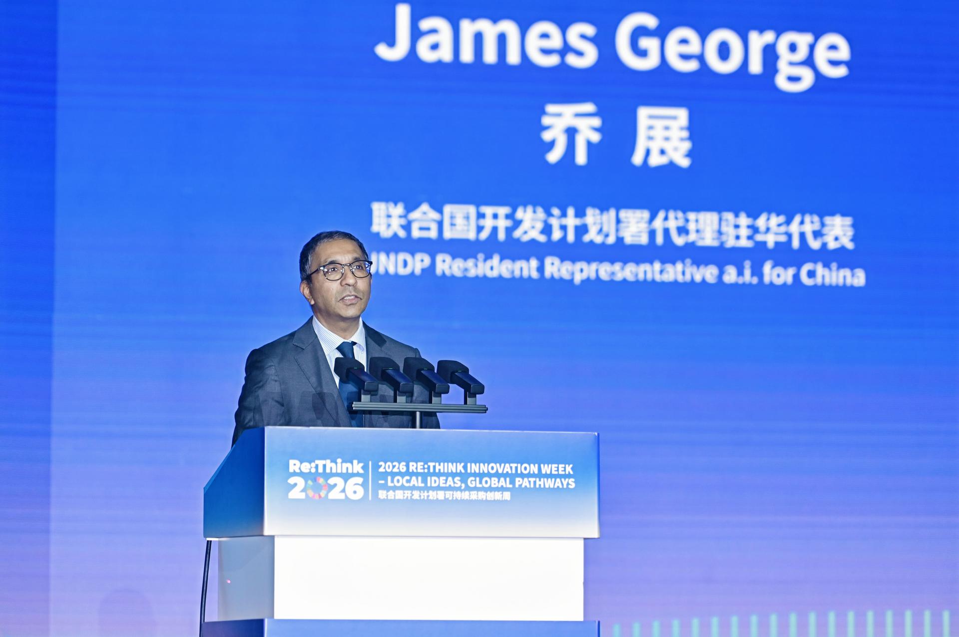Photograph of a man in a suit at a podium on a blue conference stage, James George on screen.