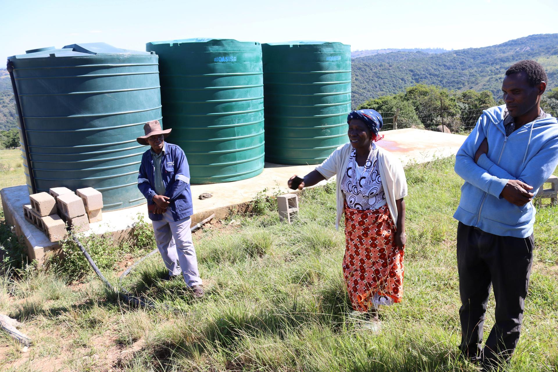 Photograph: three people walking in a grassy field beside large green water tanks.
