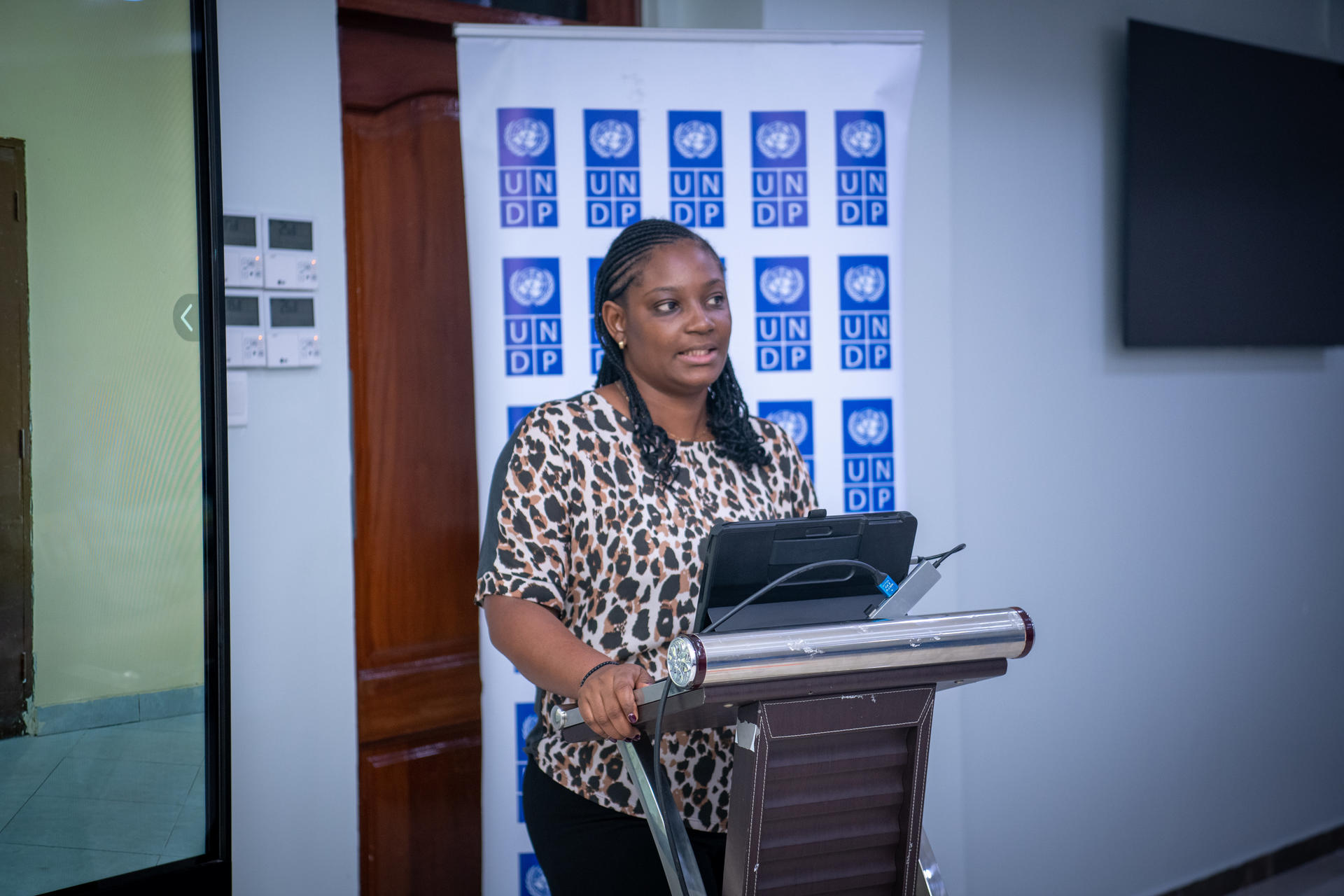 Presenter at a wooden podium with a laptop, in a conference room with a blue patterned backdrop.