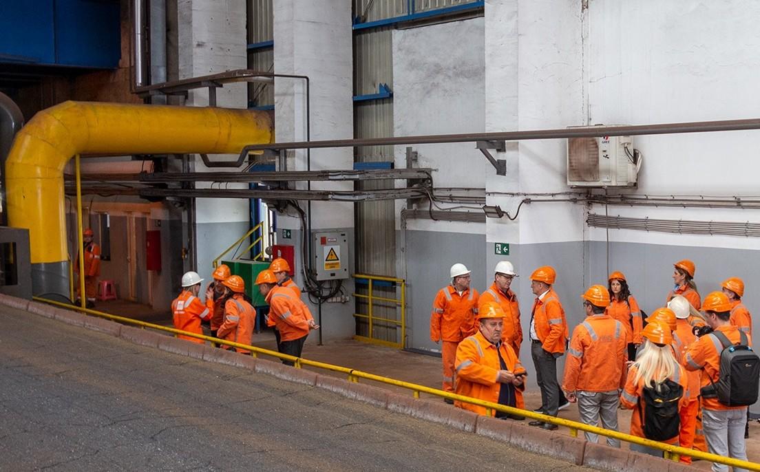 Group of workers in orange safety outfits with hard hats near yellow pipes at an industrial plant.