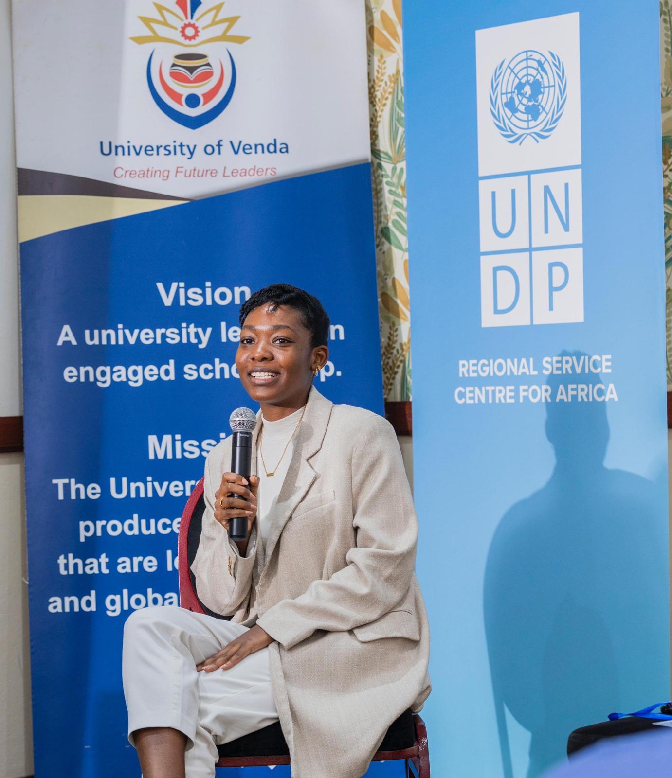 Beige-suited speaker with a microphone, face blurred, seated beside UNDP and University banners.