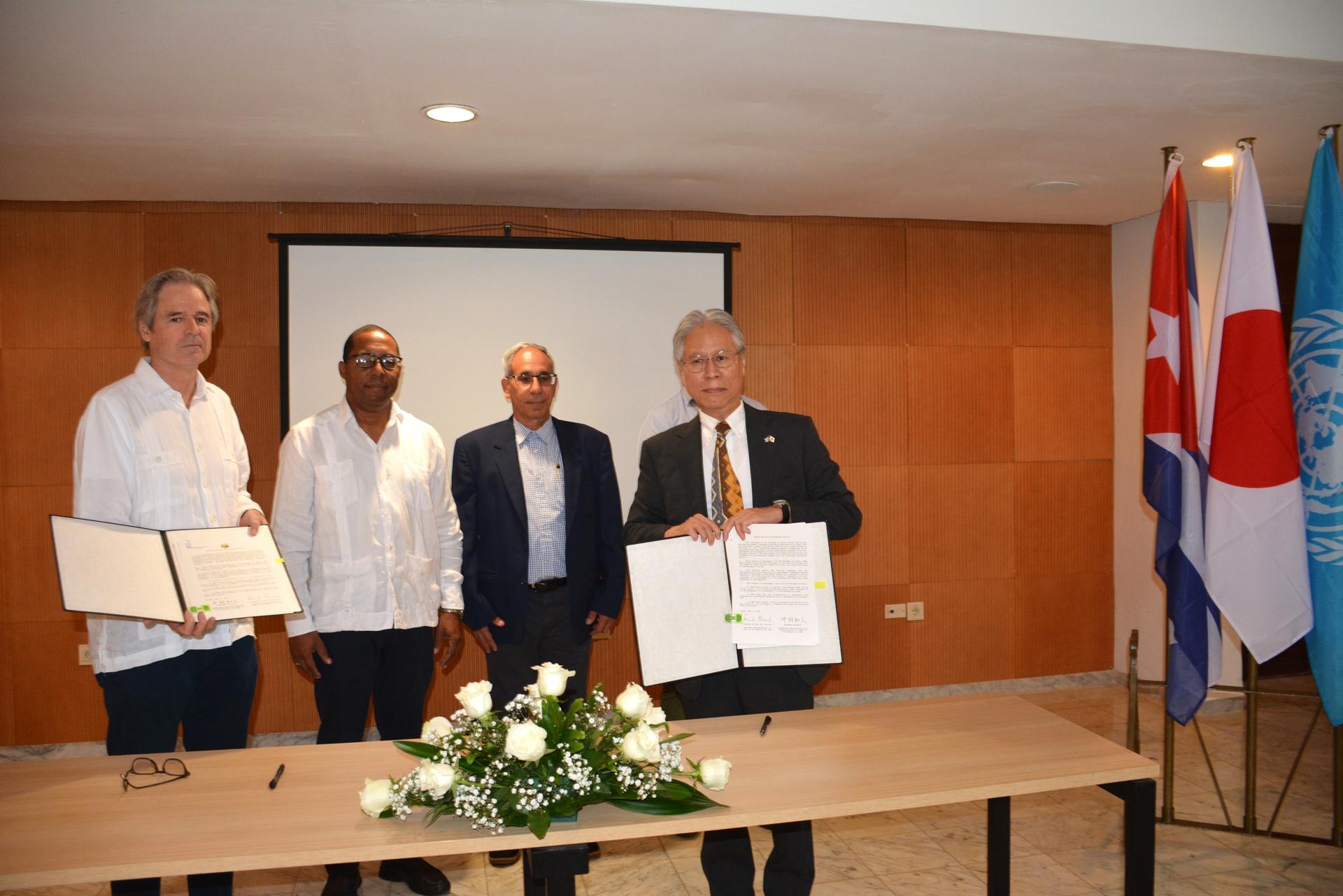 Four people stand behind a table with documents; a flower arrangement and flags in a formal room.