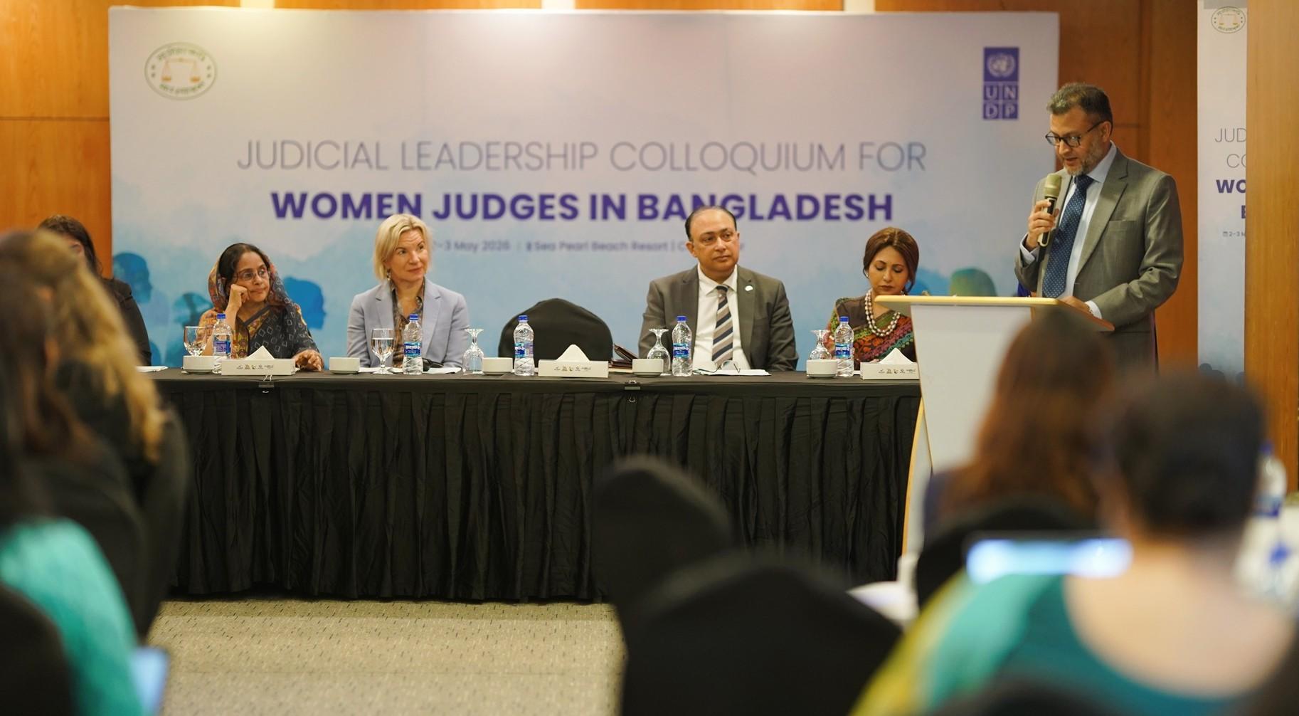 Panel of women judges at a conference table; man speaks from a podium, banner in background.