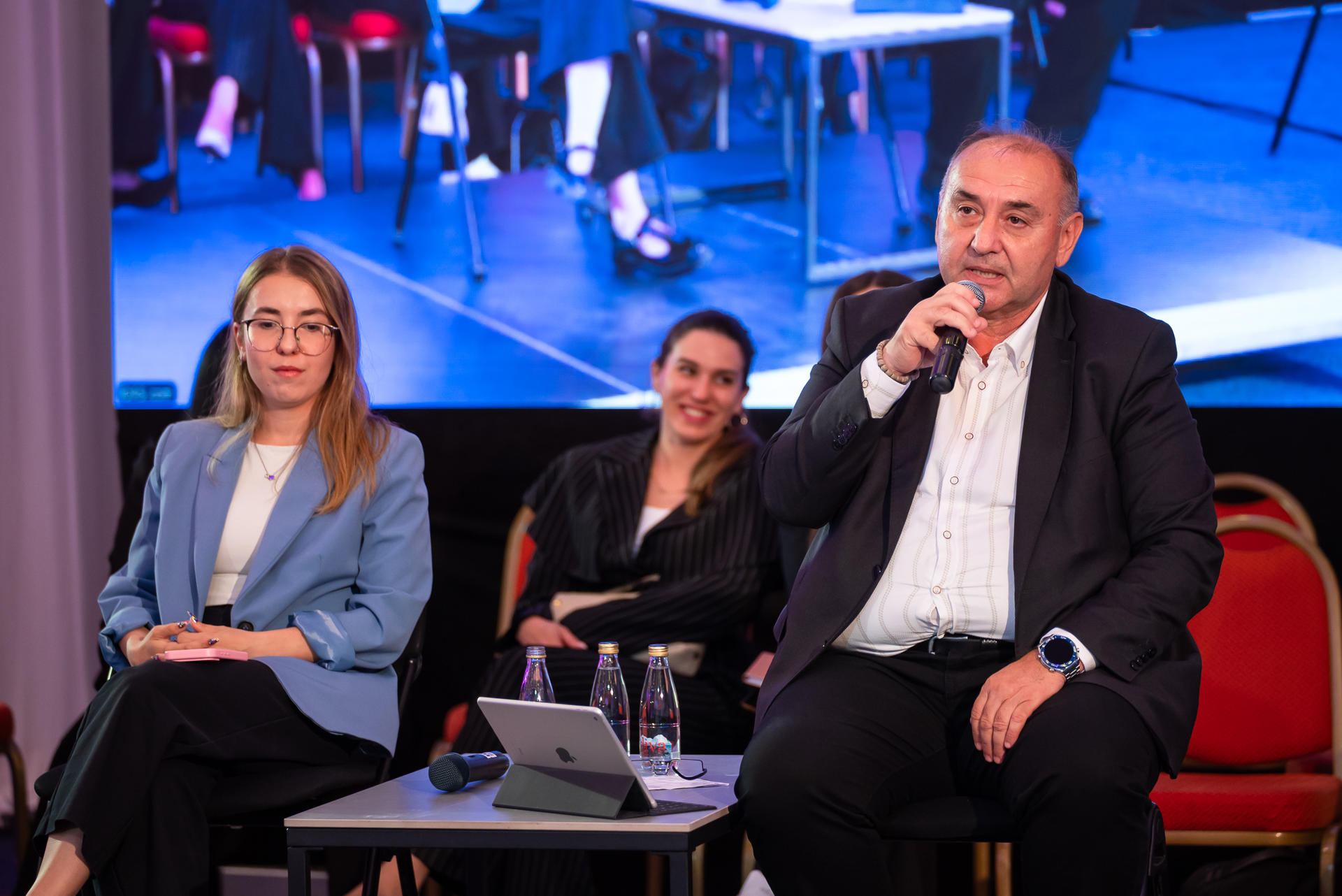 Photograph of three panelists on stage: two women and a man with microphone, blue backdrop.