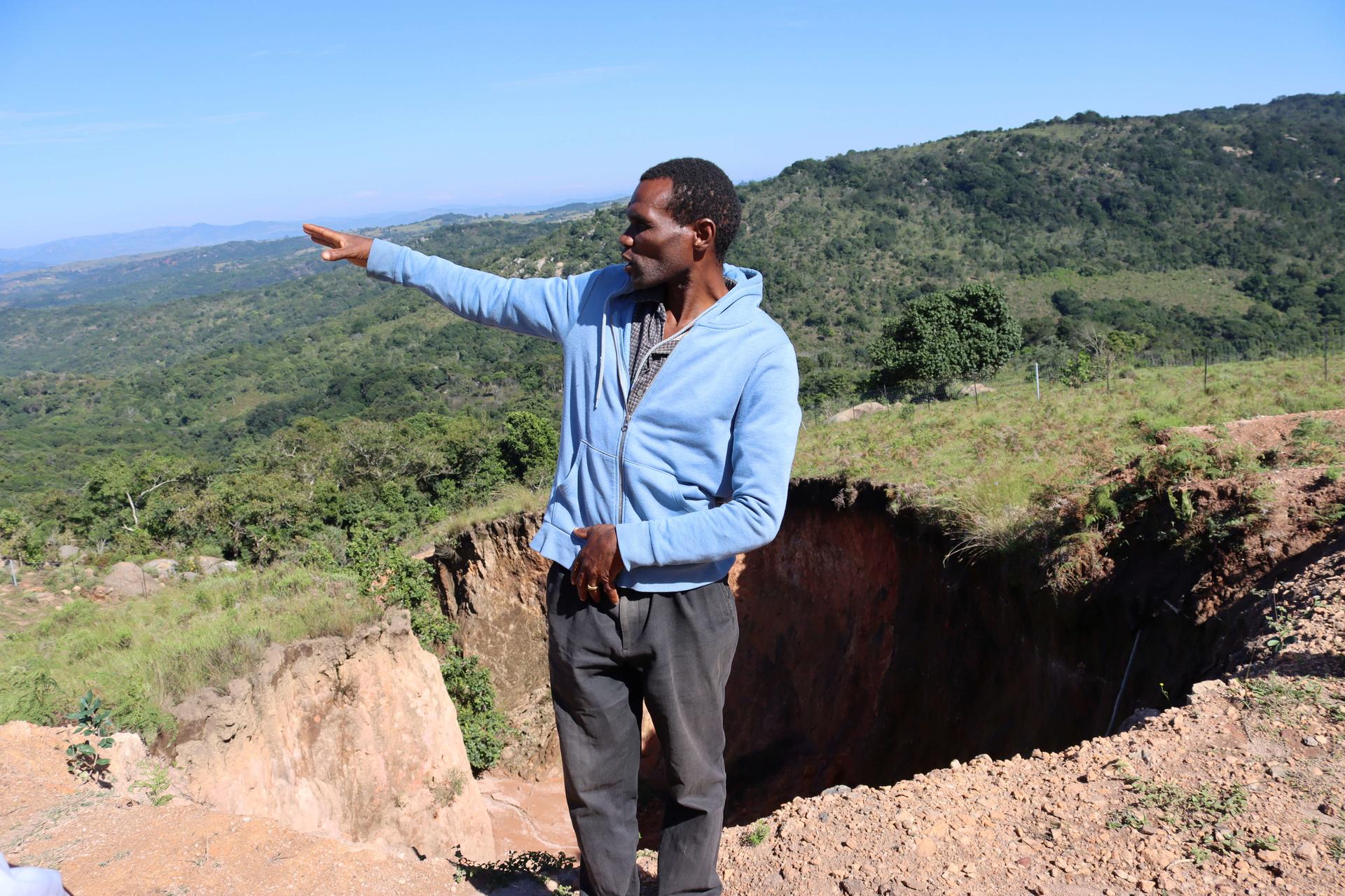 Man in blue shirt points toward a crater on rocky ground, with green hills and blue sky behind.
