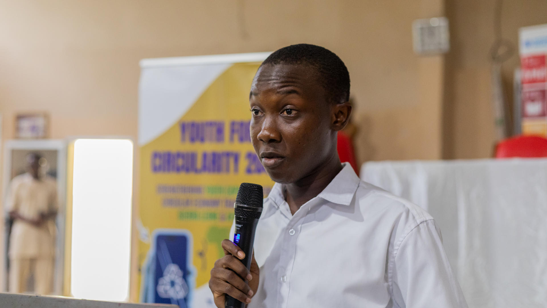 Man in a white shirt speaks into a handheld microphone at a conference, banners behind.